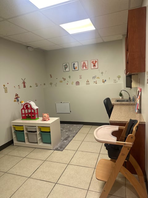 Pediatric exam room with child-sized furniture, storage bins, wall decorations, and medical equipment including chair and sink