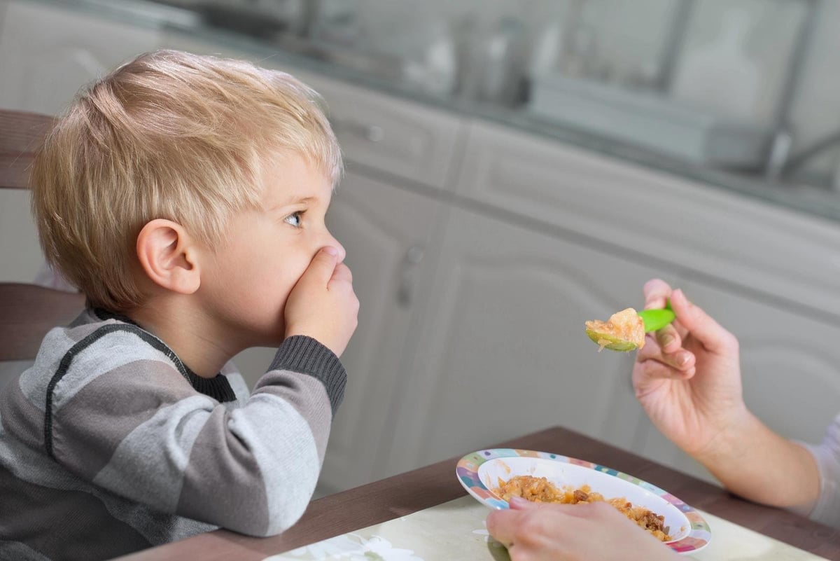 Toddler boy sitting at table looking reluctant while being offered broccoli on a plate with other food
