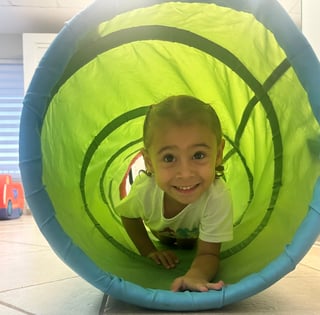 Young child smiling inside a bright green and blue tunnel toy at an indoor play facility