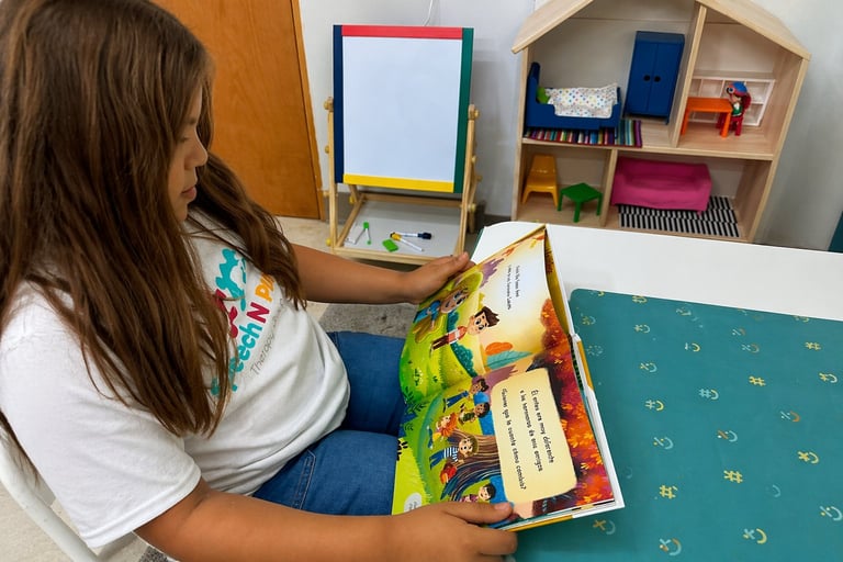 Child with long brown hair reading a colorful childrens book at a table with art supplies and shelving in the background