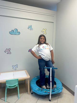 Girl standing on blue trampoline in therapy room with puzzle-themed wall decorations and small desk with chair