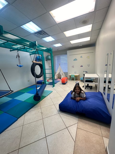 Child relaxing on a blue bean bag in a sensory room with climbing equipment, swing, and therapy tools