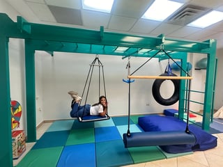 Child on hanging swing in indoor sensory play area with colorful foam floor mat and suspended equipment