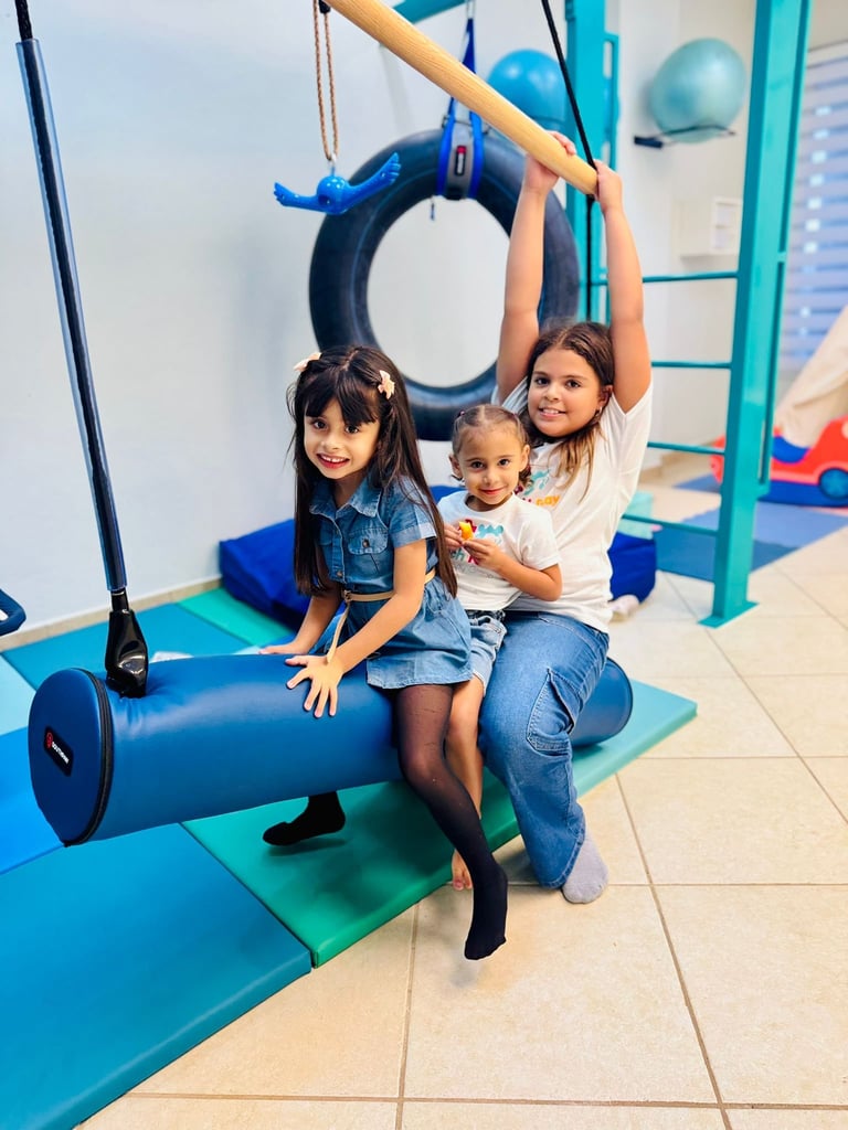 Three children playing on colorful gym equipment including a tire swing and tunnel in an indoor playground