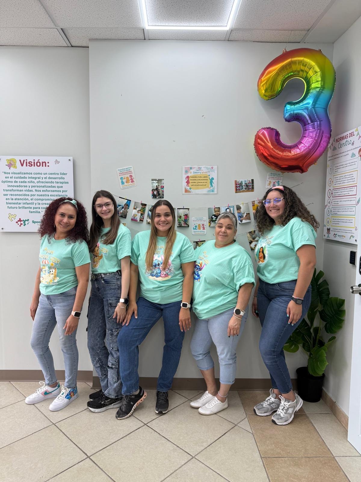 Five young women in matching mint green t-shirts standing together under a rainbow number 3 balloon in an office hallway